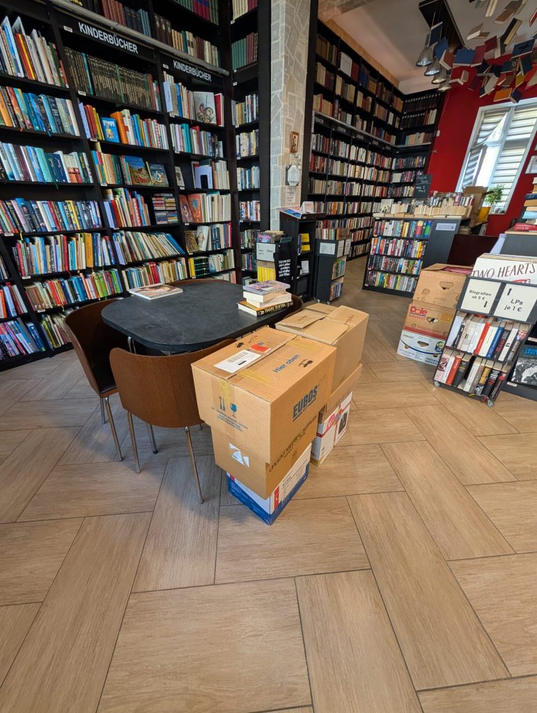 Bookstore interior with tall black shelves full of books, a round table in front, and cardboard boxes in the foreground.