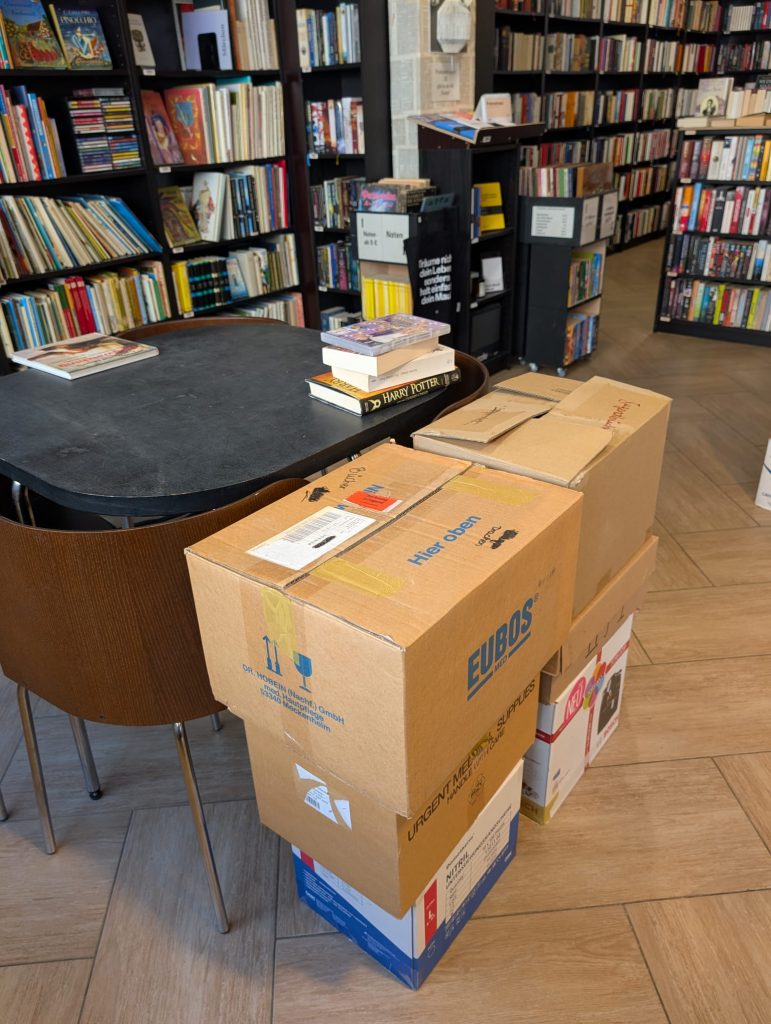 Boxed packages stacked beside a round library table in a bookstore, with tall shelves of books in the background.